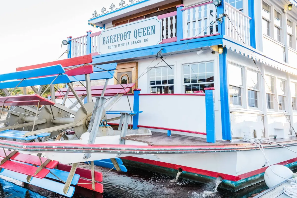 The image showcases the colorful exterior and paddle wheel of the 'Barefoot Queen' riverboat, with clear signage visible. The boat is docked, and the playful, relaxed mood is highlighted by vibrant colors and open deck space, inviting families to imagine themselves boarding for an adventure or dinner cruise.