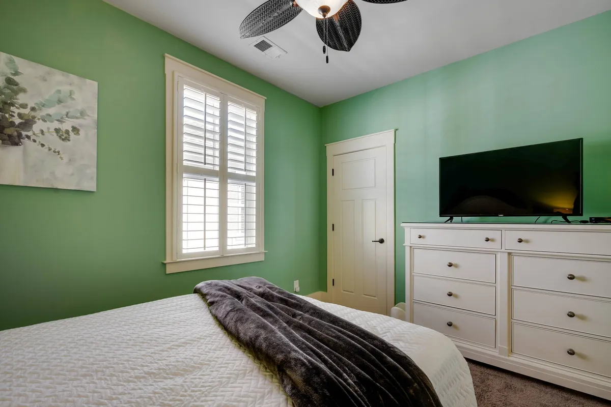 A tranquil bedroom featuring soothing green walls, white furniture, a comfortable bed with a soft blanket, and a wall-mounted TV. Natural light fills the room, enhancing the peaceful, cozy atmosphere.