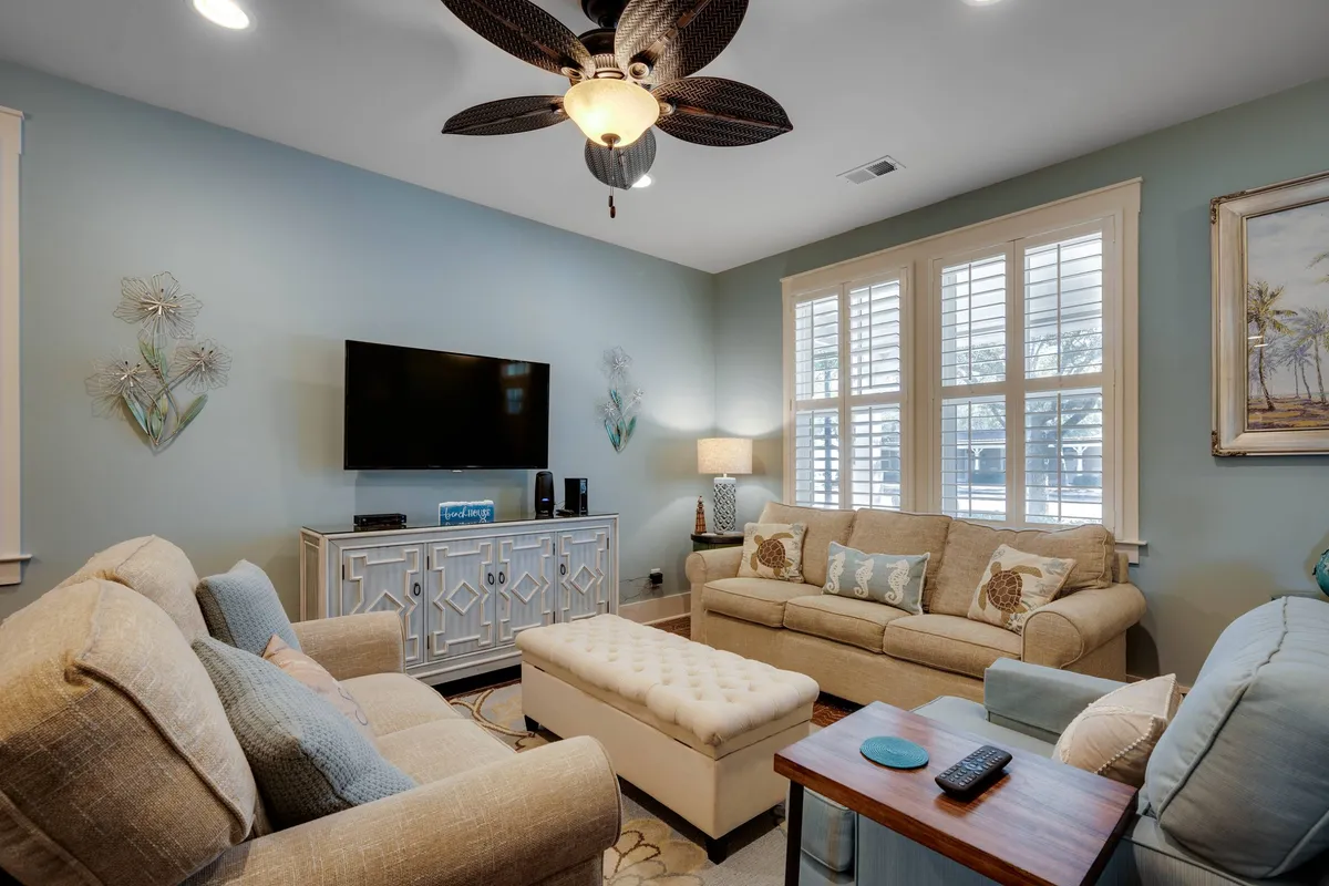 The image showcases a bright and welcoming living room with plush, comfortable sofas arranged around a stylish decorative ottoman. A large TV unit is positioned for easy viewing, and tasteful artwork adorns the walls. The space exudes warmth and is perfect for gathering with friends or family to relax, watch movies, or enjoy game nights.