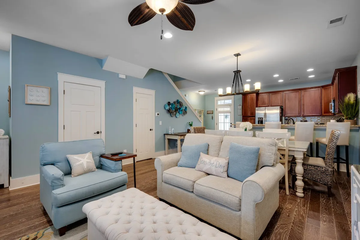 This image captures a cozy living room setup with a plush sofa adorned with decorative pillows, a charming blue armchair, and a textured ottoman. The kitchen area is visible in the background, providing a clear sense of the open-concept layout. The room's inviting and warm ambiance suggests comfort and togetherness, ideal for family gatherings or relaxing with friends.