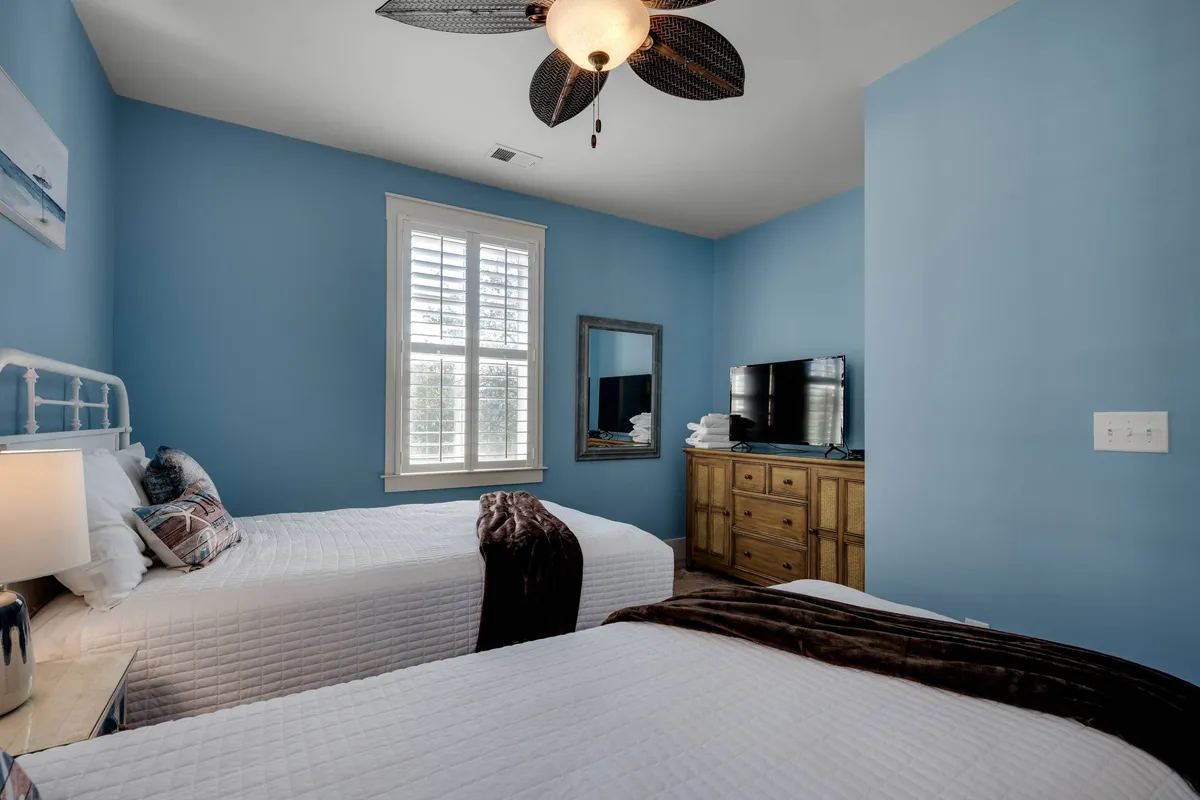 A guest bedroom with two twin beds, ceiling fan, large window with shutters, a wooden dresser, mirror, and a visible TV. The space feels calm, comfortable, and well-lit, ideal for kids, teens, or friends.
