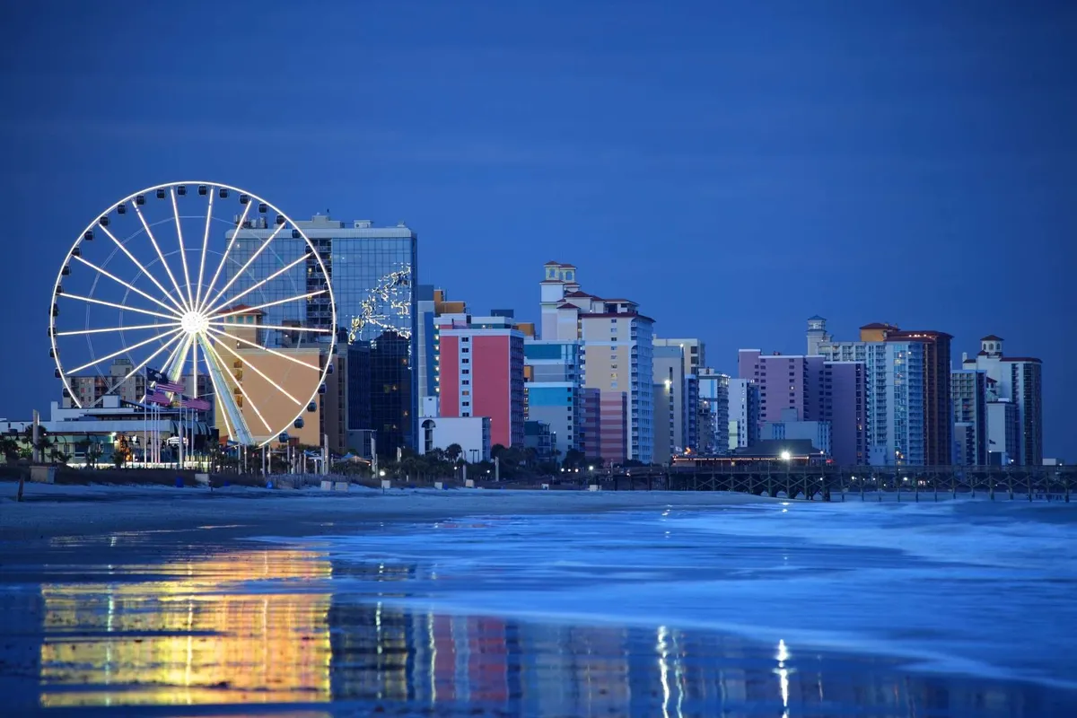 The image captures a festive oceanfront scene with a Ferris wheel, colorful buildings, and reflections of the ocean at dusk. This visually dynamic shot conveys a fun, exciting atmosphere, evoking the energy of amusement rides and nighttime entertainment near the beach.