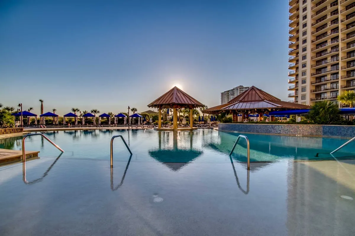 This image captures a serene swimming pool surrounded by palm trees and thatched-roof cabanas at sunset. The scene includes people relaxing by the pool, enjoying drinks, and socializing in the outdoor resort setting, evoking the ambiance of a resort getaway.