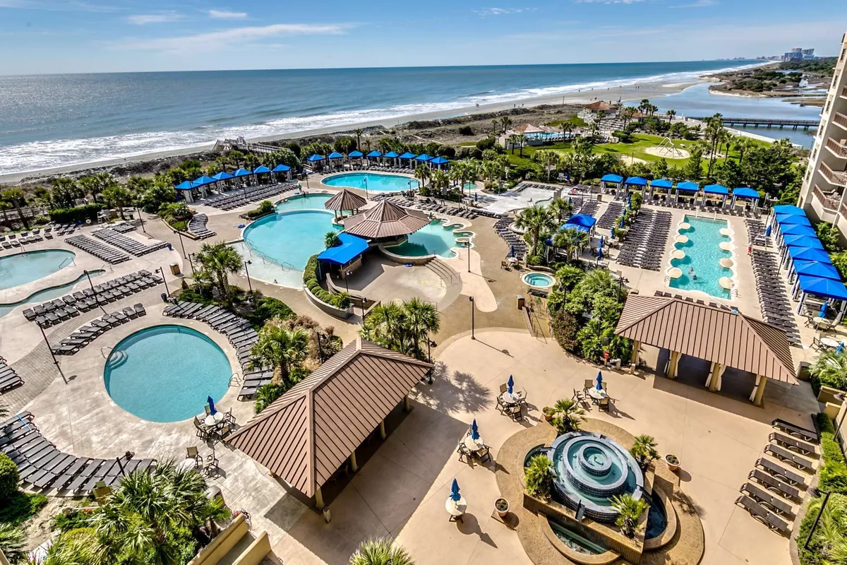 A vibrant exterior view of resort-style swimming pools lined with lounge chairs, beachfront access, landscaped gardens, and water features. Guests are seen relaxing and socializing by the pool, evoking a vacation resort atmosphere.